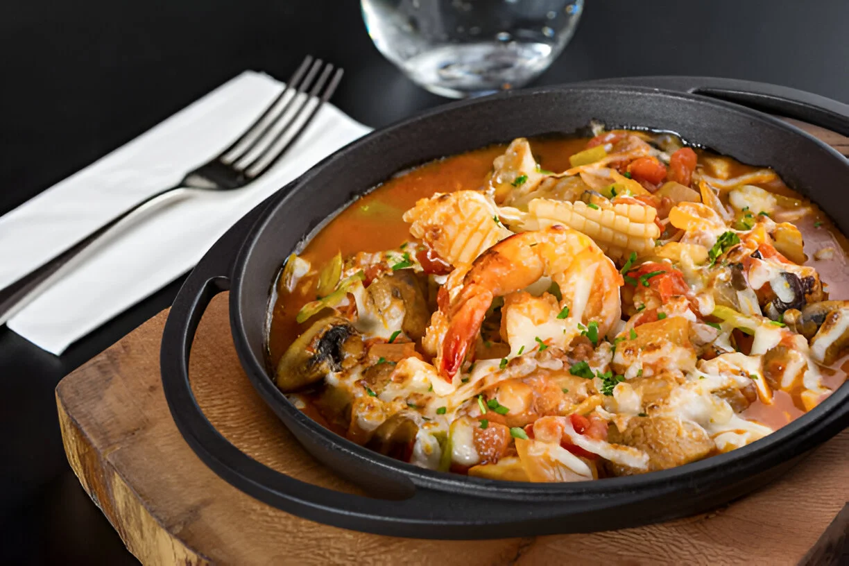 A steaming bowl of seafood gumbo filled with shrimp, crab, andouille sausage, and vegetables, served over rice with a side of crusty bread.